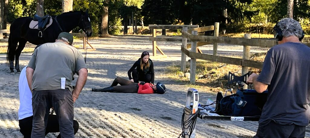 Rider on ground with bystander kneeling and horse in background, while being filmed